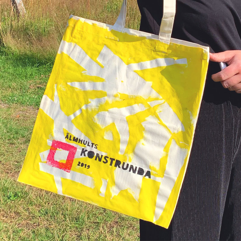 Tygväska med handtryckt gult abstrakt mönster och Älmhults konstrunda 2019 logga